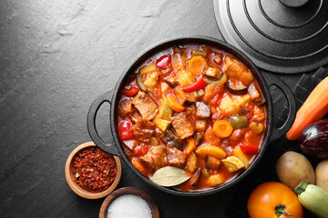 Delicious stew with vegetables in pot and ingredients on gray textured table, flat lay. Space for...