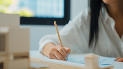 Closeup of beautiful young architect engineer hand drawing, drafting blueprint with blueprint and house model placed on working table at modern office. Focus on hand. Blurring background. Immaculate.