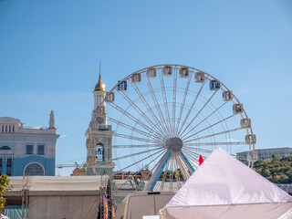 Modern observation wheel and ancient church bell tower with bright blue cloudless sky in the background in Kontraktova square, Podil, Kyiv city. Travelling and entertainment in old city center.