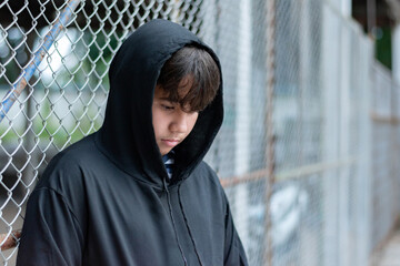 Asian youngster wearing black hoody and standing beside a metal fence in a local detention center, looking sad and wanting his freedom, broken home, lonliness, depression of young people concept.
