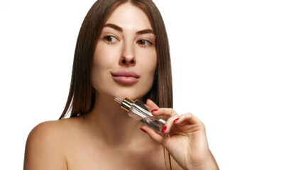 Fototapeta premium Pretty woman with bare shoulders holding serum bottle to her face for skincare against white background. Facial care treatment. Concept of beauty, healthcare, freshness and wellness, confidence. Ad