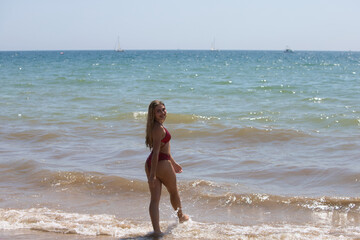 Fototapeta premium Teenage, Latin, blonde, young woman is having fun on the beach shore playing with the waves. The girl is wearing a metallic red bikini and is on vacation in andalusia, Spain.