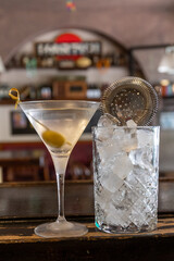 close up shot of a martini cocktail with olive in the typical martini glass, on the wooden counter of a bar with tumbler and strainer. Adult beverage, leisure, relaxation mood, italian aperitif.