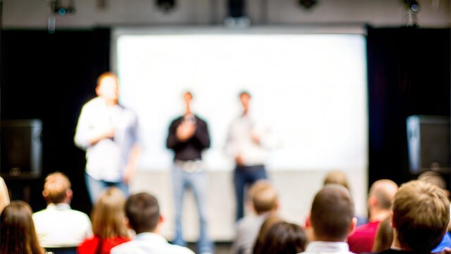 A blurred image of a startup pitch event, with an audience watching presenters on stage.

