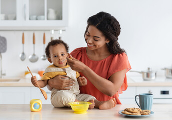 Baby Weaning. Caring Black Mother Feeding Little Toddler Son From Spoon In Kitchen, Loving African American Mommy Giving Porridge Or Mash Fruit Puree To Cute Little Child At Home, Closeup Shot