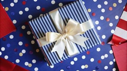 Obraz premium A Navy Blue and White Striped Gift Box with a Creamy White Bow on a Blue Background with White, Red, and Gold Confetti