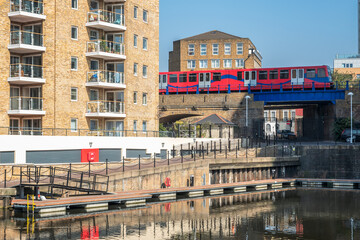 A Docklands Light Railway (DLR) train passes over a bridge near modern apartment buildings and a calm waterfront at Limehouse Basin in London, UK