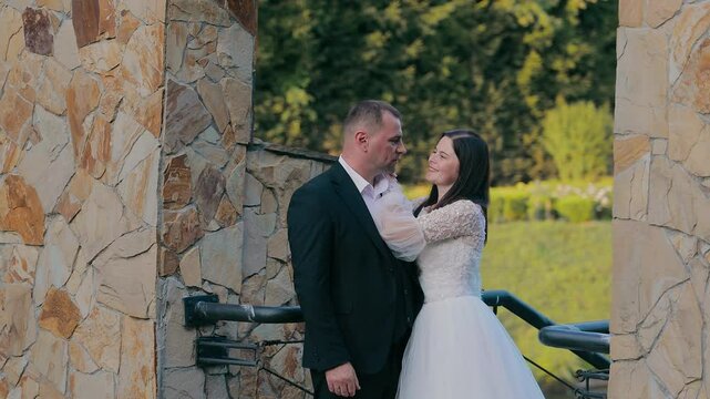Bride and Groom Sharing Tender Moment by Stone Wall, A bride and groom stand close by a rustic stone wall, sharing a tender moment as she gently touches his lapel