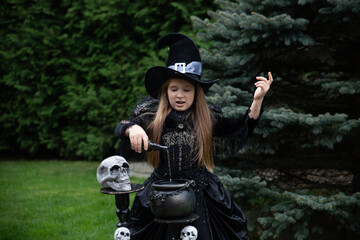 Charming sorceress European girl 8 years old against the background of  tree in a black dress pours green liquid into the cauldron Halloween potion skull hand up