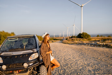 A young woman takes a break, leaning against her rugged off-road vehicle, sipping a hot drink while...