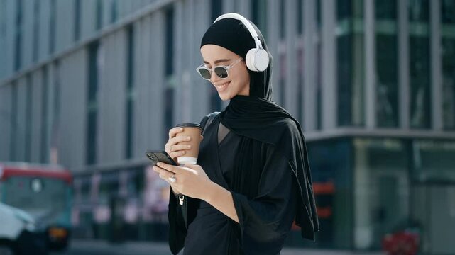 Video of beautiful woman with hijab listening to music with headphones while using smartphone walking throught the city