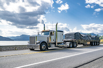 Classic American idol big rig white semi truck with chrome parts transporting covered cargo on flat...