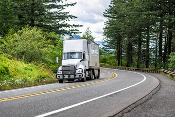 Day cab white big rig semi truck with huge roof spoiler and protection grille guard transporting cargo in dry van semi trailer running on the turning mountain road with forest on the sides