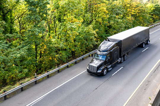 Stylish black big rig semi truck with extended cab transporting cargo in conestoga flat bed railer running on the autumn highway road with forest on the side