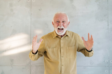A senior Caucasian businessman stands indoors, hands raised in an astonished gesture, dressed in...