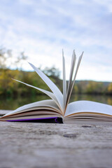 Open book with pages flipping in the wind. Reading on the lake wooden dock on autumn day. Learning enjoying tranquility of nature. Peaceful outdoor environment. Selective focus. Blurred background.