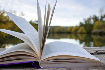 Open book with pages flipping in the wind. Reading on the lake wooden dock on autumn day. Learning enjoying tranquility of nature. Peaceful outdoor environment. Selective focus. Blurred background.