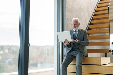 A focused senior Caucasian male in a tailored suit uses a laptop while sitting on wooden stairs in a well-lit, modern office environment.