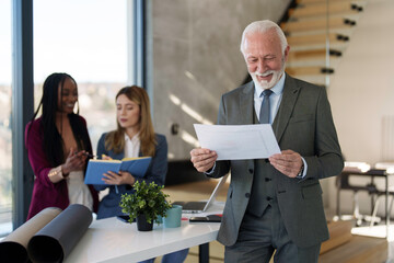 A smiling senior Caucasian businessman examines a paper in a well-lit office, with young female colleagues in the background, one of diverse ethnicity, all in professional attire.
