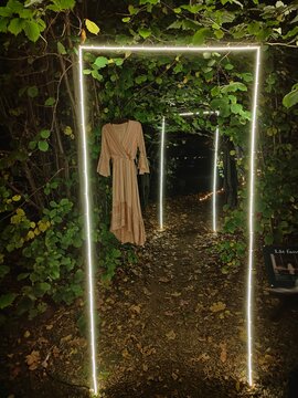 A mysterious outdoor installation with illuminated frames and a hanging dress, set amidst leafy foliage and a dimly lit path, creating an enchanting and ethereal atmosphere
