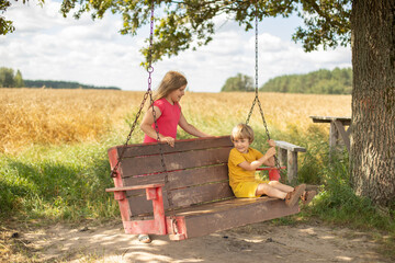 A blond 5 year old boy and a blond 8 year old girl. Brother and sister swing on a swing on a hot summer day in the shade of a large oak tree in a meadow, with a large yellow field of wheat in the back