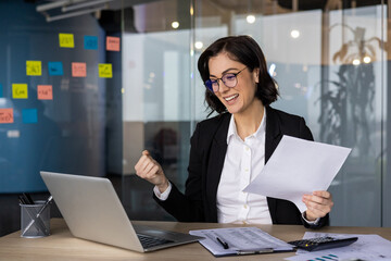 Young businesswoman celebrating success working on laptop at office desk. Holding documents,...