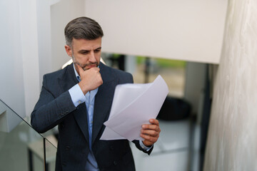 A pensive mid adult Caucasian businessman reviews papers, wearing a suit in a well-lit, modern office, embodying professionalism and concentration.