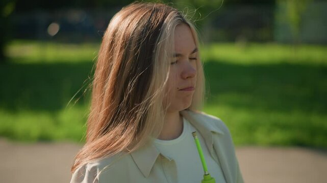Young woman stands in bright sunlight, cooling her face with a green air pump, squinting slightly as gentle breeze flows through her hair, sunlight filters through trees and a building in background
