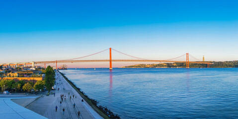 Panoramic view of 25 de abril bridge over tagus river in lisbon at sunset, with clear blue sky and christ the king statue in background, people enjoying waterfront promenade