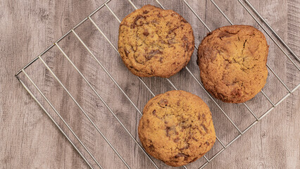 Three freshly baked chocolate chip cookies on a cooling rack over rustic wood. Ideal for dessert or snack themes.