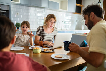 Fototapeta premium Mother working on laptop while family has breakfast in kitchen