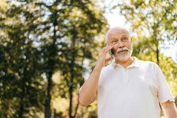 Low-angle view of elderly male looking peaceful and happy chatting on mobile phone in sunny summer...