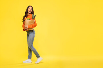 Full length shot of young European woman with grocery in paper bag after shopping in supermarket, standing over yellow background and looking back at free space