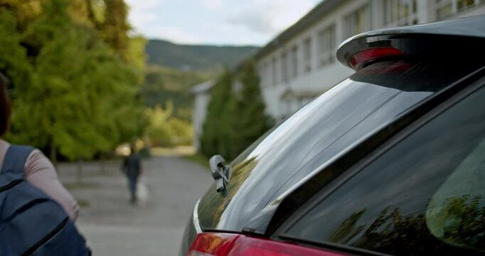 A young teenage girl with a backpack walks towards school after getting dropped off by her parent. She walks away from the car towards the school building.