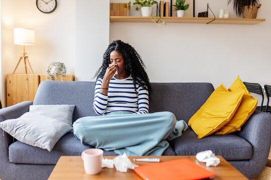 Young African woman is blowing her nose while sitting on a sofa, covered with a blanket, feeling unwell at home