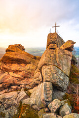 At sunset, the Oresnik granite rock formation glows warmly, showcasing a wooden summit cross atop the lookout point. The surrounding Jizera Mountains offer stunning panoramic views.
