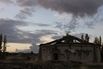 old abandoned dovecote with a partially collapsed roof, set against a dramatic evening sky. The surrounding grass and distant buildings add a rural charm, highlighting the beauty of decay and solitude
