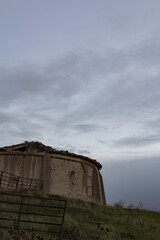 old abandoned dovecote with a partially collapsed roof, set against a dramatic evening sky. The surrounding grass and distant buildings add a rural charm, highlighting the beauty of decay and solitude