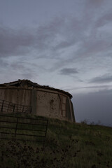 old abandoned dovecote with a partially collapsed roof, set against a dramatic evening sky. The surrounding grass and distant buildings add a rural charm, highlighting the beauty of decay and solitude