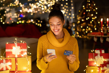 Joyful shopper lady buying Christmas gifts online, using smartphone and credit card, sitting in decorated with lights room. New year home shopping