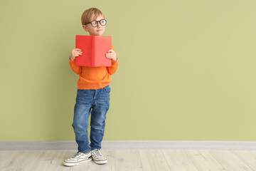 Cute little boy in eyeglasses with open book standing against color wall