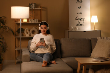 Young woman in headphones chatting with mobile phone on cozy sofa in living room at night