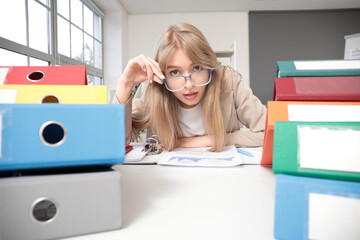 Young female accountant in eyeglasses at workplace with stacks of folders in office