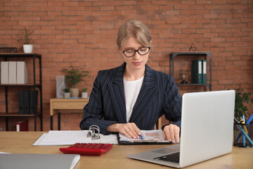 Young female accountant in eyeglasses doing paper work at workplace with laptop and calculator in...