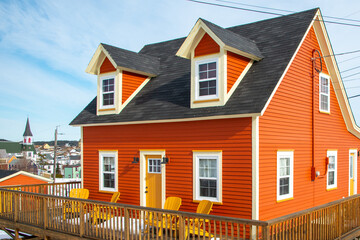 The exterior corner of a vintage bright orange wooden cottage with white trim, double hung windows, peaked roof and gabled windows.The doors are colorful yellow. The yard is enclosed with a wood fence