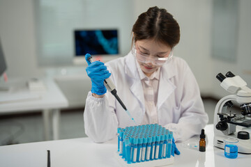 Young female scientist wearing lab coat and gloves using micropipette dropping blue liquid into test tubes doing research in modern laboratory with microscope