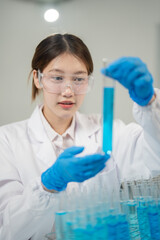 Fototapeta premium Young scientist wearing lab coat and gloves carefully examining test tube filled with blue liquid, conducting research or experiment in modern laboratory setting