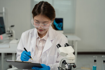 Asian female scientist wearing lab coat and protective glasses writing notes during experiment in modern laboratory with microscope and computer