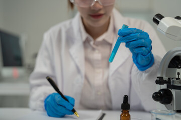 Scientist wearing lab coat and gloves analyzing blue solution in test tube and taking notes while conducting research in laboratory with microscope