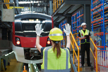 Female traffic controller in safety vest jacket with green hard hat gives signal to the driver of electric locomotives red train in the high tech maintenance plant. © AU USAnakul